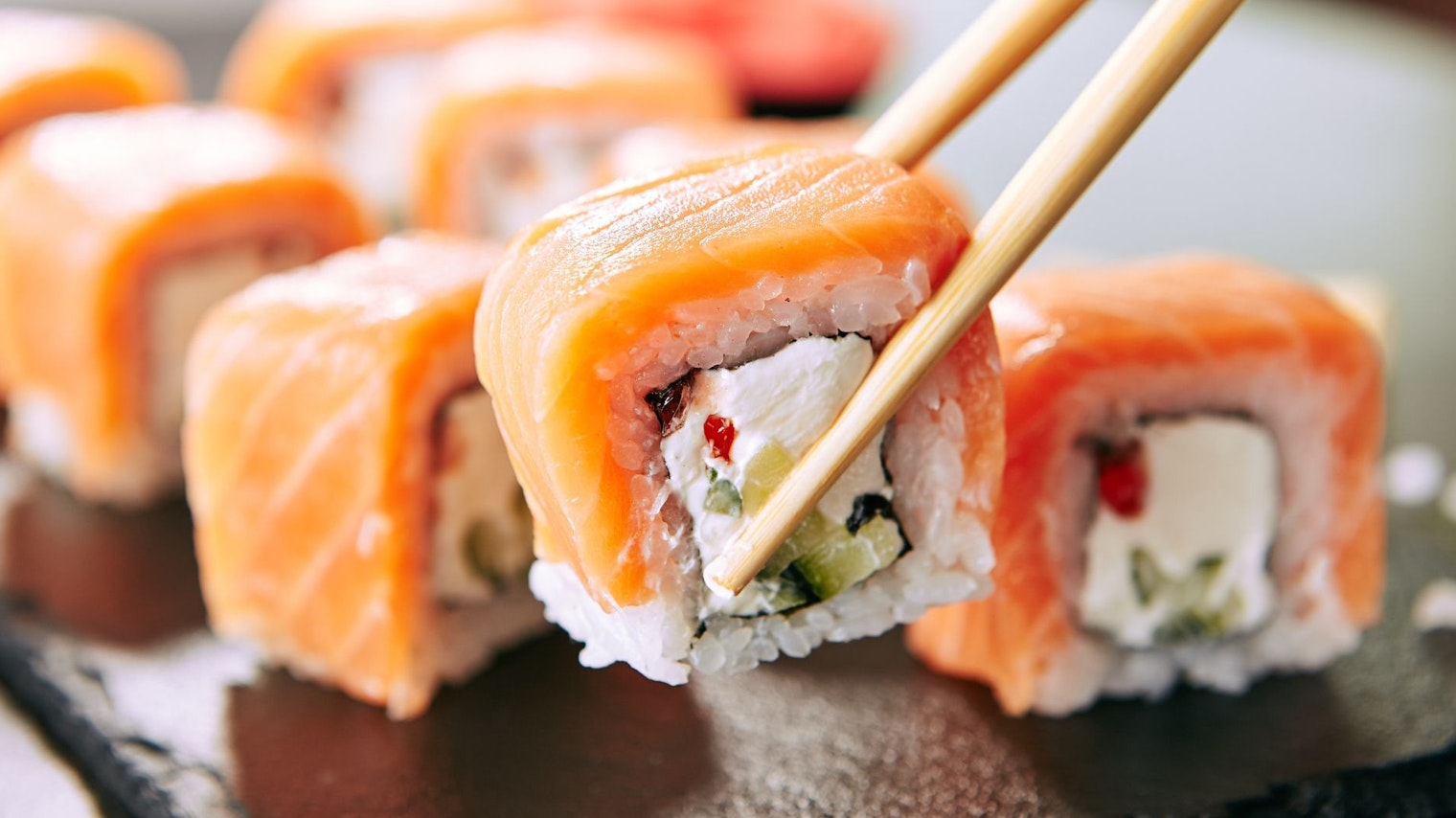 Chopstick holding Sushi Rolls Set with Salmon and Cream Cheese and Cuccumber on Black Slate Plate Close Up. Uramaki, Nori Maki or Futomaki Sushi with Trout Fillets, Soy Sauce and Wasabi