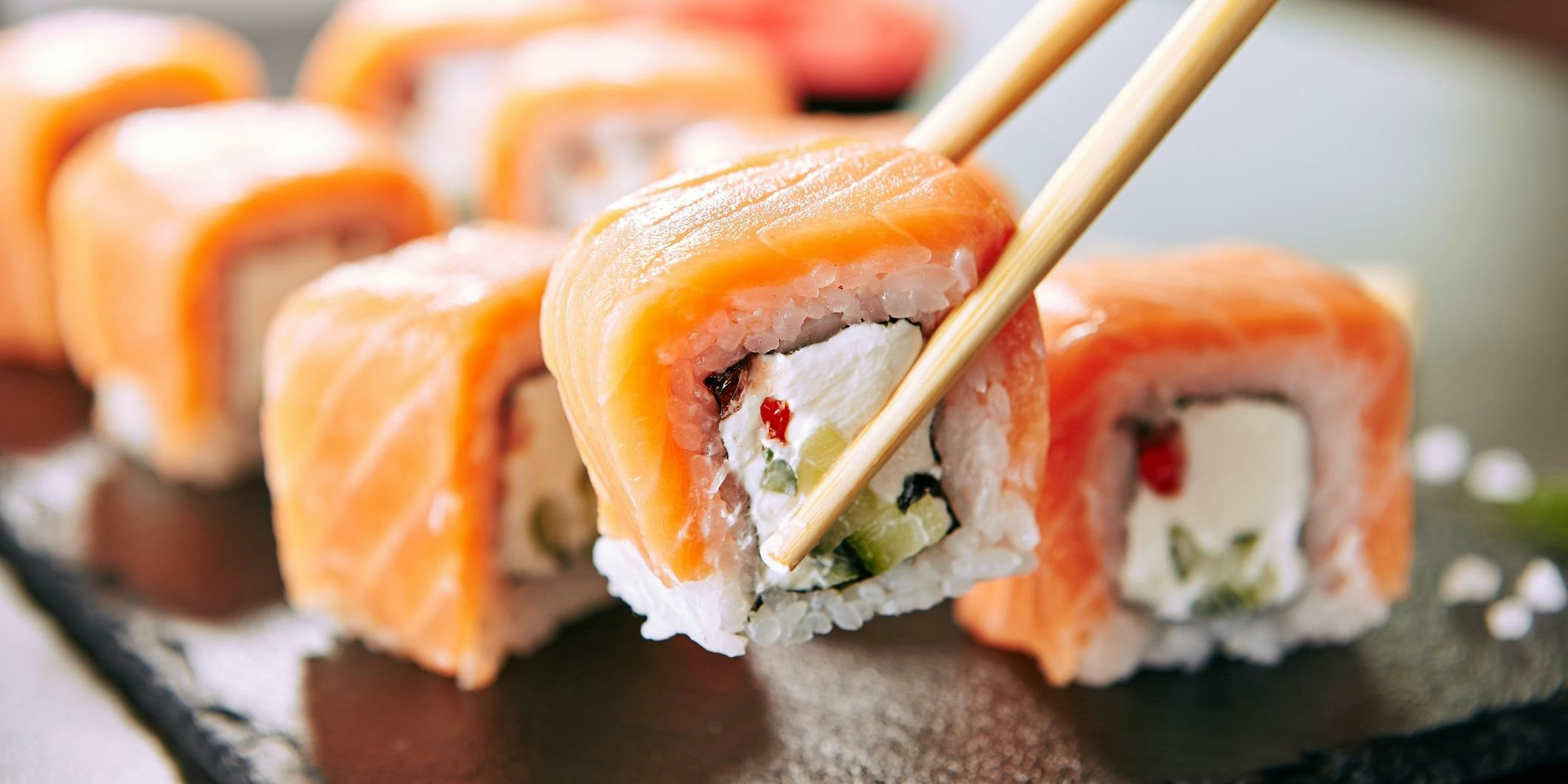Chopstick holding Sushi Rolls Set with Salmon and Cream Cheese and Cuccumber on Black Slate Plate Close Up. Uramaki, Nori Maki or Futomaki Sushi with Trout Fillets, Soy Sauce and Wasabi