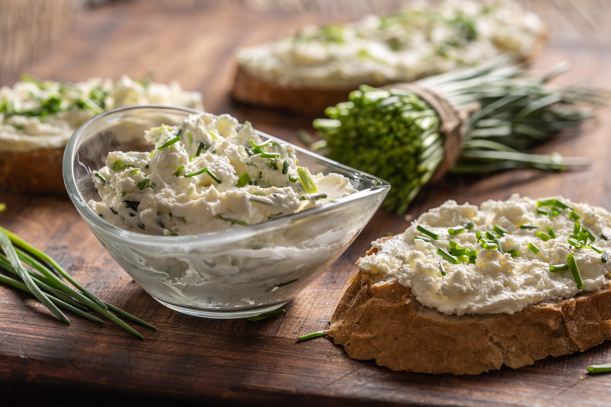 A bowl of homemade cream cheese spread with chopped chives surrounded by bread slices with spread and a bunch of freshly cut chives.